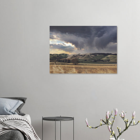 Landscape artwork of Te Hauke hills under dark clouds with beams of light breaking through.