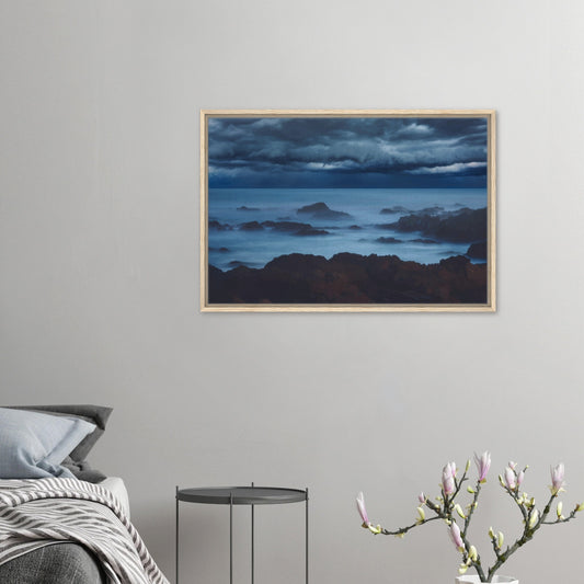 Deep navy and steel-blue tones sweep across a moody sky, while waves crash and swirl around jagged rocks, softened by a misty, dreamlike haze.