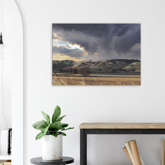 Landscape artwork of Te Hauke hills under dark clouds with beams of light breaking through.