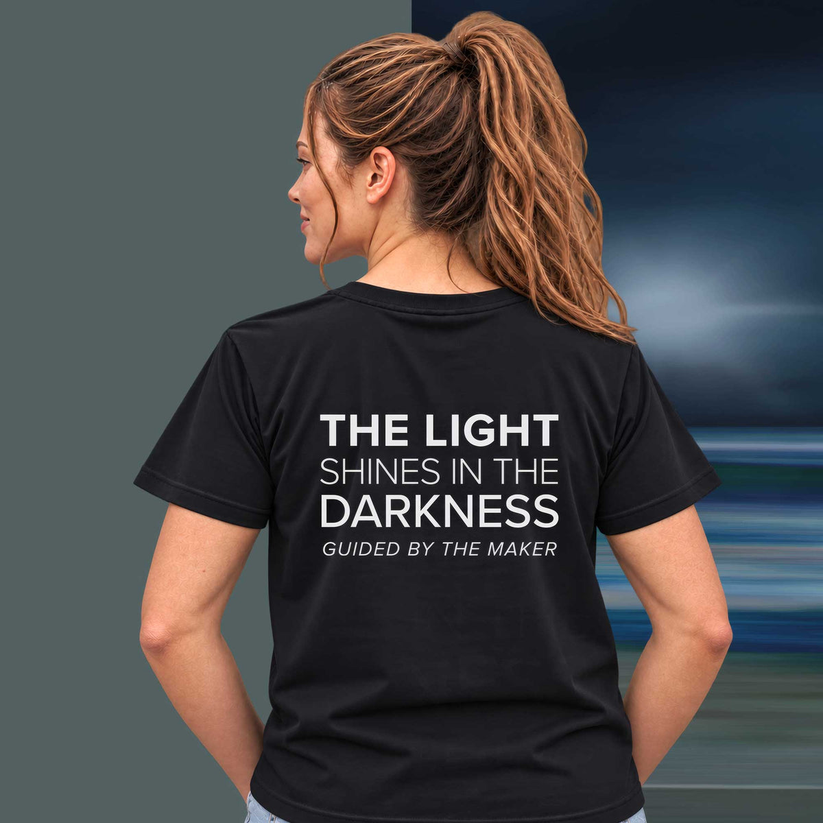 Model wearing the 'The light shines in the darkness' back print tee in black, large message printed on the back - Blend Designs NZ