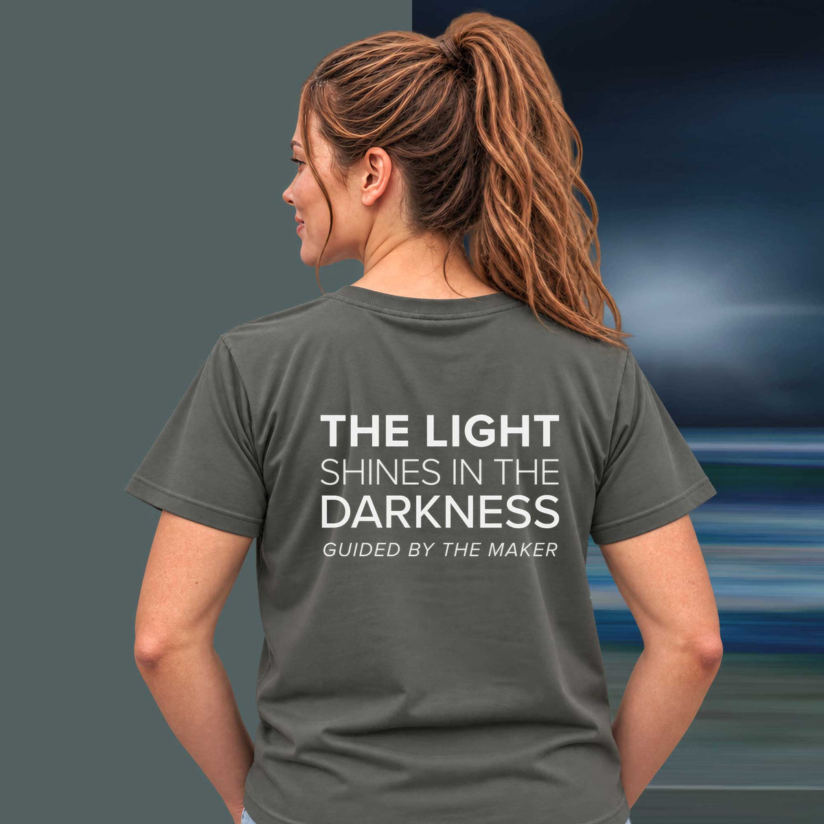 Model wearing the 'The light shines in the darkness' back print tee in charcoal, large message printed on the back - Blend Designs NZ