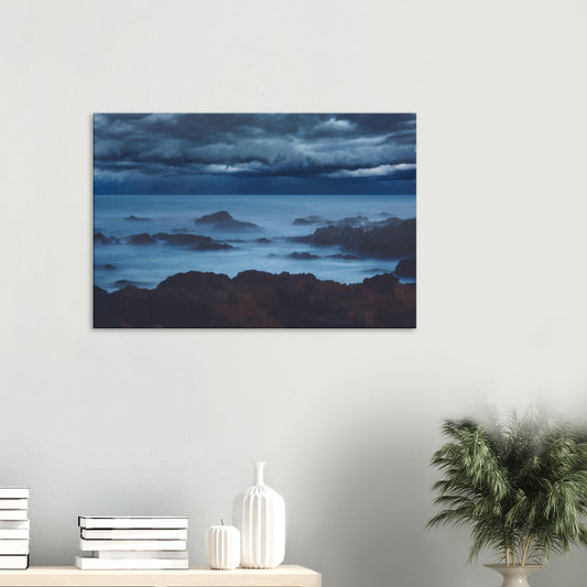 Deep navy and steel-blue tones sweep across a moody sky, while waves crash and swirl around jagged rocks, softened by a misty, dreamlike haze.